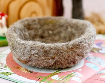 May include: A small, round, brown felted bowl. The bowl is sitting on a colorful book with a tree and other decorative elements in the background. The bowl has a textured, natural appearance.