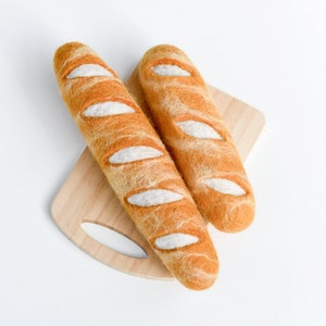 May include: Two orange and white felt baguettes sit on a light brown wooden chopping board. The baguettes have white, oval details and a textured appearance, suggesting a handmade craft item. The background is plain white.