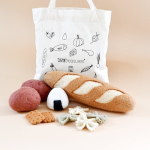 Könnte beinhalten: Eine weiße Stofftasche mit einer schwarz-weißen Illustration verschiedener Lebensmittel, darunter ein Fisch, eine Birne, ein Kürbis, eine Karotte, ein Baguette, eine Erdbeere, ein Stück Käse, ein Karton Milch, Erbsen und eine Zwiebel. Die Tasche ist gefüllt mit Filz-Lebensmitteln, darunter ein Baguette, zwei rote Kartoffeln, ein Reisball und Nudeln. Der Text "taratreasures" ist auf die Tasche gedruckt.
