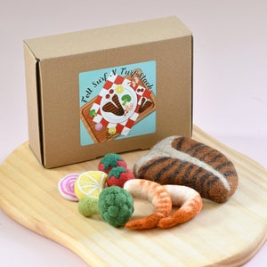 Puede incluir: Un juego de comida de fieltro con una caja de cartón y una pegatina que dice "Fell Surf N Turf Stack". El juego incluye filete de fieltro, camarones, tomates, limón, brócoli y una tabla de cortar de madera.