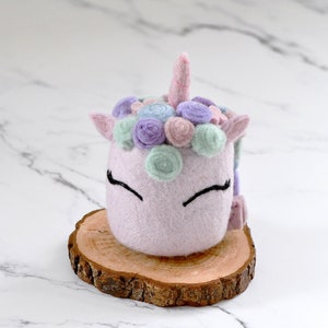 May include: A pink felt unicorn with a pastel flower crown, closed eyes, and a pink horn. The unicorn is sitting on a wooden slice.