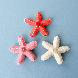 Puede incluir: Tres estrellas de mar de fieltro en diferentes colores: rojo, rosa y crema. Cada estrella de mar tiene puntos blancos en su superficie.