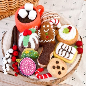 Puede incluir: Una colección de alimentos de fieltro, que incluye una taza de chocolate caliente con malvaviscos, un hombre de jengibre, un higo y varios pasteles. Los artículos están dispuestos sobre una rodaja de madera, con una cesta en el fondo.