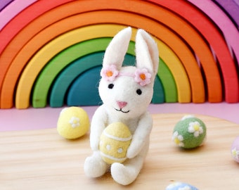 May include: A white felt bunny rabbit holding a yellow felt Easter egg with pink felt flowers on its head. The bunny is sitting on a wooden surface with other felt Easter eggs in blue, green, and yellow.