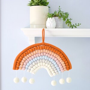May include: A felt rainbow wall hanging with a light orange, peach, pink, light blue, and white color scheme. The rainbow has a loop for hanging and white pom-poms hanging from the bottom.