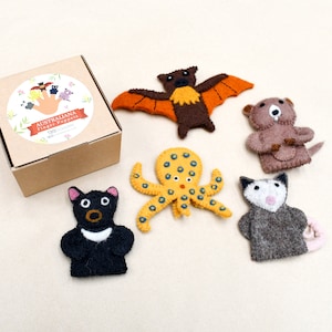 Puede incluir: Un conjunto de títeres de dedo de fieltro con animales australianos. Los títeres incluyen un murciélago, un pulpo, un wombat, un zarigüeya y un demonio de Tasmania. Los títeres están empaquetados en una caja marrón con el texto "AUSTRALIANA Finger Puppets" impreso en la parte superior.