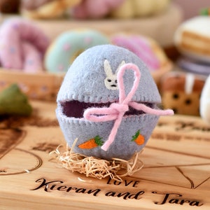 May include: A pale blue felt Easter egg, decorated with a pink bow, a white bunny, and orange carrot appliqués. The egg is open, showing a purple interior, and rests on straw. The wooden board below has the engraved text "Kieran and Tara".