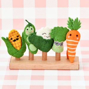May include: A wooden display stand holding five felt vegetable figures: corn, peas, cauliflower, broccoli, and a carrot. Each vegetable has a smiling face and is crafted with bright colors. The background is a pink and white checkered pattern.