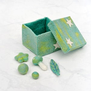 May include: A teal green felt box with a lid decorated with white stars. The box is open and there are several felt items inside, including a mushroom, a leaf, a star, and two balls.