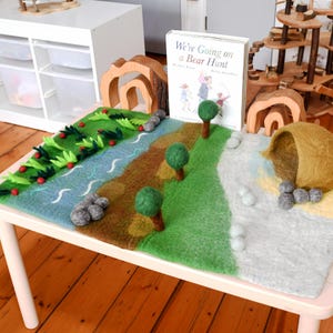 May include: A felt play mat featuring a landscape with a blue river, green grass, and brown earth. The mat includes three-dimensional felt trees, gray stone accents, and a brown felt cave. A copy of the book We're Going on a Bear Hunt is placed upright on the mat, accompanied by two wooden arch structures.