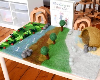 May include: A felt play mat featuring a landscape with a blue river, green grass, and brown earth. The mat includes three-dimensional felt trees, gray stone accents, and a brown felt cave. A copy of the book We're Going on a Bear Hunt is placed upright on the mat, accompanied by two wooden arch structures.