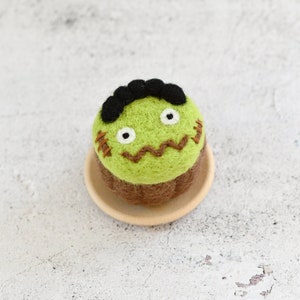 May include: A green felt cupcake with a stitched-on face, black hair, and brown icing. The cupcake is sitting on a small wooden plate.