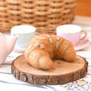 May include: A felt croissant with a brown and beige color scheme, sitting on a wooden slice. The croissant is decorated with small, beige felt circles.