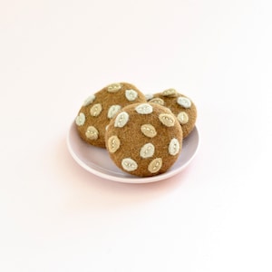 Puede incluir: Tres galletas de fieltro marrón con detalles blancos cosidos, dispuestas sobre un plato pequeño de color rosa claro. Las galletas tienen un aspecto texturizado y un patrón de detalles blancos espaciados. El fondo es rosa liso y suave.