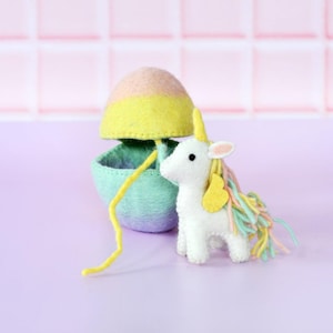 May include: A white felt unicorn with a yellow horn and colorful mane stands next to a felt egg-shaped container. The egg is yellow, pink, and green. A yellow string connects the unicorn and the container. The background is a light purple.