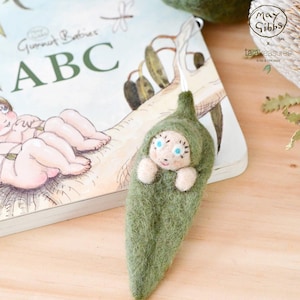 Può includere: Una bambola di feltro di un bambino avvolto in un bozzolo di feltro verde. La bambola ha gli occhi azzurri e un viso bianco. La bambola è seduta su un libro con le parole "Gumboot Babies ABC" sulla copertina.