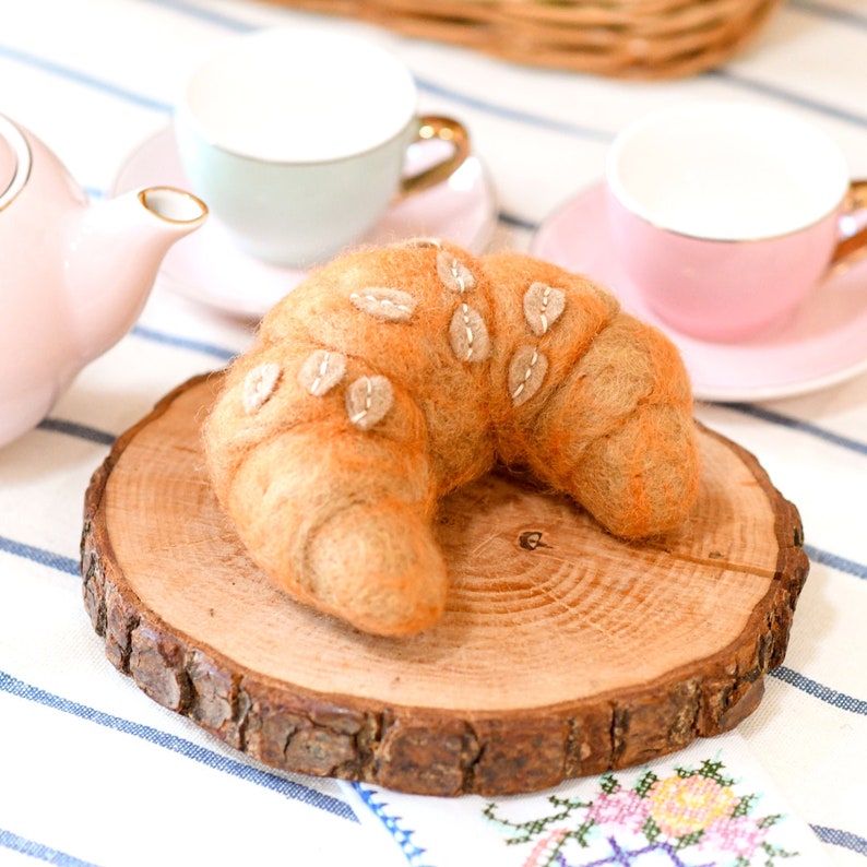 May include: A felt croissant with a brown and orange color scheme, decorated with white felt leaves, sits on a wooden slice. The croissant is on a white and blue striped tablecloth.