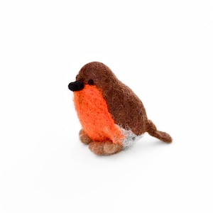 Könnte beinhalten: Eine kleine, handgefilzte Rotkehlchen-Figur mit braunem Kopf und Rücken, orangefarbener Brust und weißem Bauch. Das Rotkehlchen sitzt auf einer weißen Oberfläche.