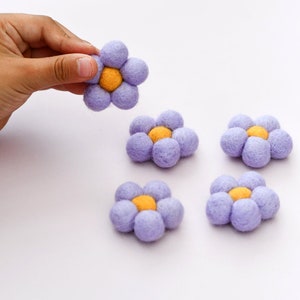 May include: Five purple felt flowers with yellow centres. The flowers are arranged in a row on a white surface.