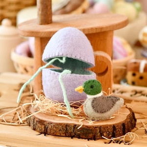 May include: A handcrafted Easter duckling and egg set. The duckling is grey, green, and yellow, placed on a wooden slice with straw. The lavender egg is open, showing a green interior. A wooden toy is in the background.
