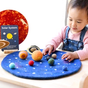 Könnte beinhalten: Ein Kind spielt mit einem Filz-Sonnensystem-Set. Das Set enthält eine blaue Filzmatte mit weißen Sternen und Planeten aus Filzkugeln. Die Planeten sind in verschiedenen Farben, darunter Rot, Orange, Gelb und Blau. Das Kind hält eine Filzkugel in der Hand. Das Set enthält auch eine Karte mit der Aufschrift "Sonnensystem".