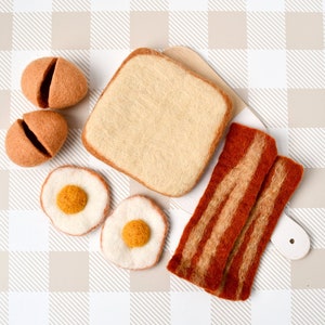 Pode incluir: Conjunto de comida de feltro com uma fatia de pão, dois ovos fritos, duas fatias de bacon e dois ovos marrons. A comida está disposta sobre uma superfície quadriculada branca e marrom.