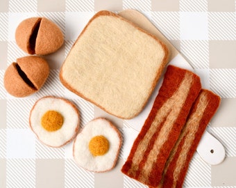 Puede incluir: Conjunto de comida de fieltro que incluye una rebanada de pan, dos huevos fritos, dos tiras de tocino y dos huevos marrones. La comida está dispuesta sobre una superficie a cuadros blancos y marrones.