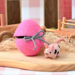 May include: A pink felt egg with a gray bow, open to reveal a dark interior, sits on a wooden board. A small felt pig with black spots stands nearby. The background includes a wooden fence and a pink barn.