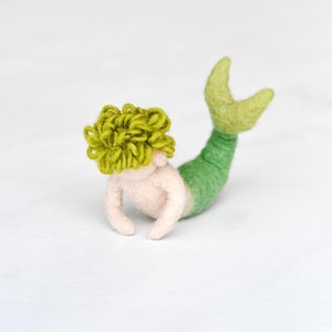 May include: A green and white felt mermaid with curly green hair. The mermaid is lying on its stomach with its tail curled up.