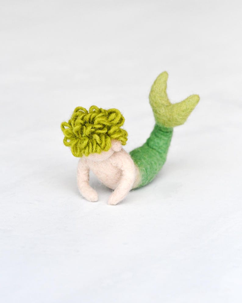 May include: A green and white felt mermaid with curly green hair. The mermaid is lying on its stomach with its tail curled up.
