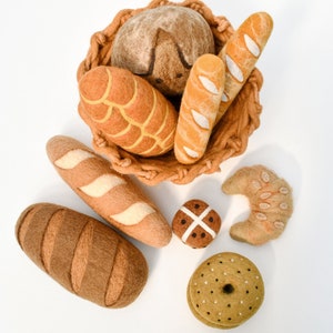 May include: A collection of felt food items, including a loaf of bread, a baguette, a croissant, a bagel, and a hot cross bun. The items are arranged in a basket and on a white surface.