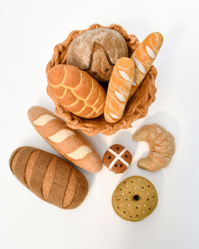 May include: A collection of felt food items, including a loaf of bread, a baguette, a croissant, a bagel, and a hot cross bun. The items are arranged in a basket and on a white surface.