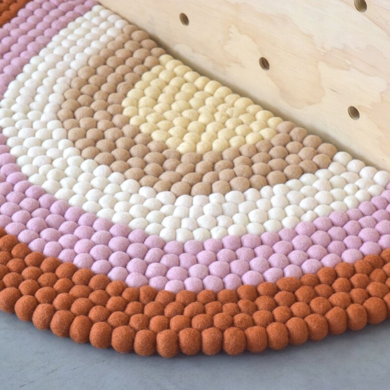 May include: A semi-circular rug with a textured surface made of small, round, felted balls. The rug features a gradient of colors, including rust, pink, white, tan, and peach. The rug is placed against a light-colored wooden surface.