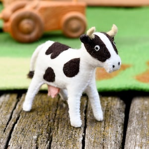 May include: A white and black felt cow toy with a pink udder. The cow is standing on a weathered wooden surface.