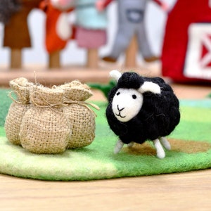 May include: A black felt sheep toy with white face and ears, standing on a green felt play mat with a brown burlap sack beside it.