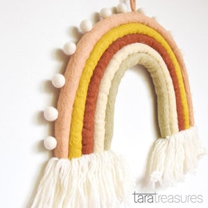 May include: A felt rainbow wall hanging with a white fringe and white pom poms. The rainbow is made of five colors: pink, yellow, orange, cream, and green.