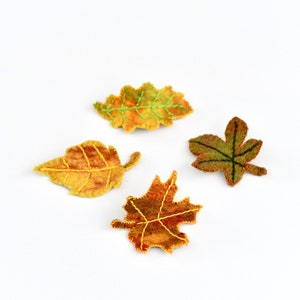 May include: Four felt leaf ornaments in various shades of green, yellow, and orange. The leaves are stitched with green thread and have a rustic, handmade look.