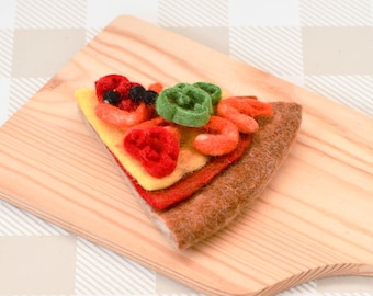 May include: A felt pizza slice with red and yellow toppings, green peppers, and black olives on a wooden cutting board.