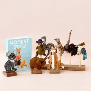 Peut inclure: Une collection de figurines d'animaux en feutre, dont un koala, un wombat, un renard et un émeu, exposée avec un livre intitulé "Wombat Stew". Les figurines sont sur des socles en bois, et le livre a une couverture bleu clair avec des illustrations.
