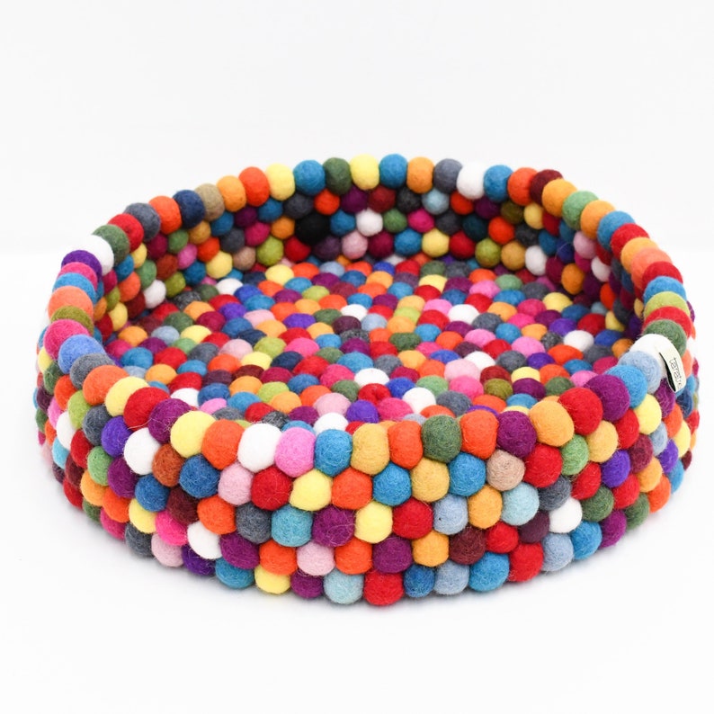 May include: A round pet bed made of colorful felt balls in various shades of red, orange, yellow, green, blue, purple, and white. The bed has a raised edge and a dense, textured surface, providing a comfortable and visually appealing space for pets.