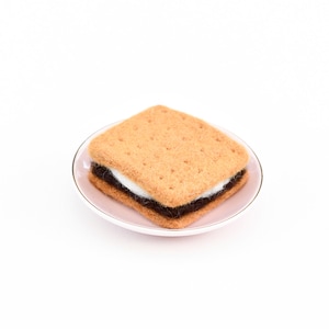 May include: A felt s'more on a pink saucer. The s'more is made of two graham cracker squares with a layer of chocolate and marshmallow in between.