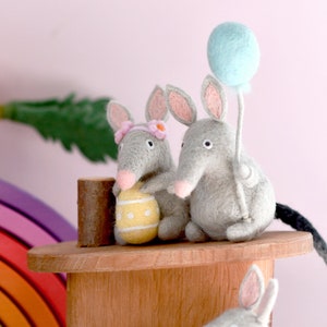 May include: Two gray felt mice with pink ears and noses, one holding a yellow Easter egg and the other holding a light blue balloon. The mice are sitting on a wooden platform with a rainbow in the background.