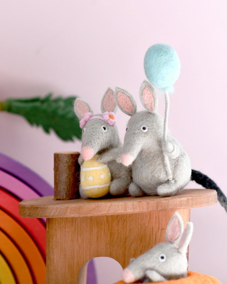 May include: Two gray felt mice with pink ears and noses, one holding a yellow Easter egg and the other holding a light blue balloon. The mice are sitting on a wooden platform with a rainbow in the background.