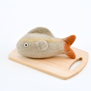 May include: A hand-felted gray fish with orange tail fins sits on a wooden cutting board. The fish has a yellow stripe and a black eye.