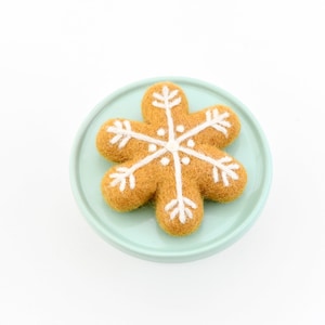 May include: A felt snowflake ornament in shades of brown and white, resting on a light blue plate. The snowflake design features white embroidered details. The ornament is a festive holiday decoration.