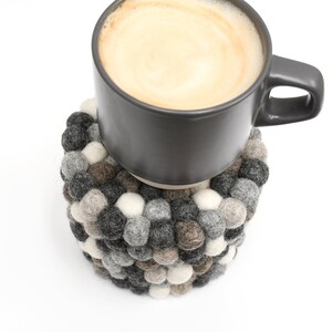 May include: A gray ceramic mug filled with coffee sits on a round felt coaster made of small, gray, white, and brown balls.