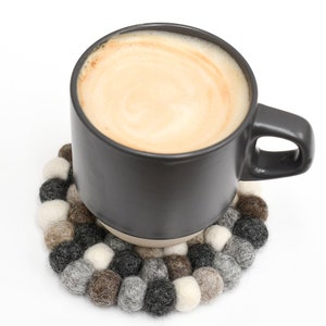 May include: A gray ceramic mug filled with coffee or tea, sitting on a round felt coaster made of brown, gray, and white wool balls.