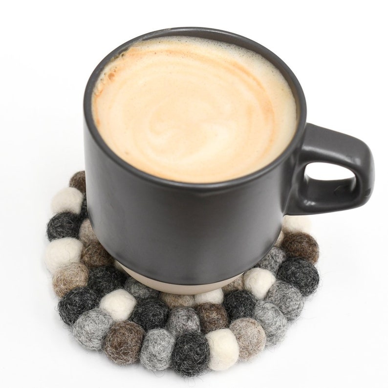 May include: A gray ceramic mug filled with coffee or tea, sitting on a round felt coaster made of brown, gray, and white wool balls.