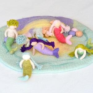 May include: A set of felt mermaid dolls on a rainbow-colored felt play mat. The mermaids are in various colors, including green, pink, purple, and blue. The play mat is a circle with a light blue border and a rainbow-colored center.