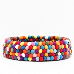 May include: A round, multi-colored felt ball basket. The basket is made of rows of colorful felt balls in shades of red, orange, yellow, green, blue, purple, and white. The basket is a vibrant and decorative item.